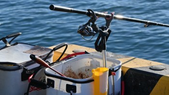A fishing rod and tackle box are set on the edge of a dock over water, with tools and bait containers nearby.