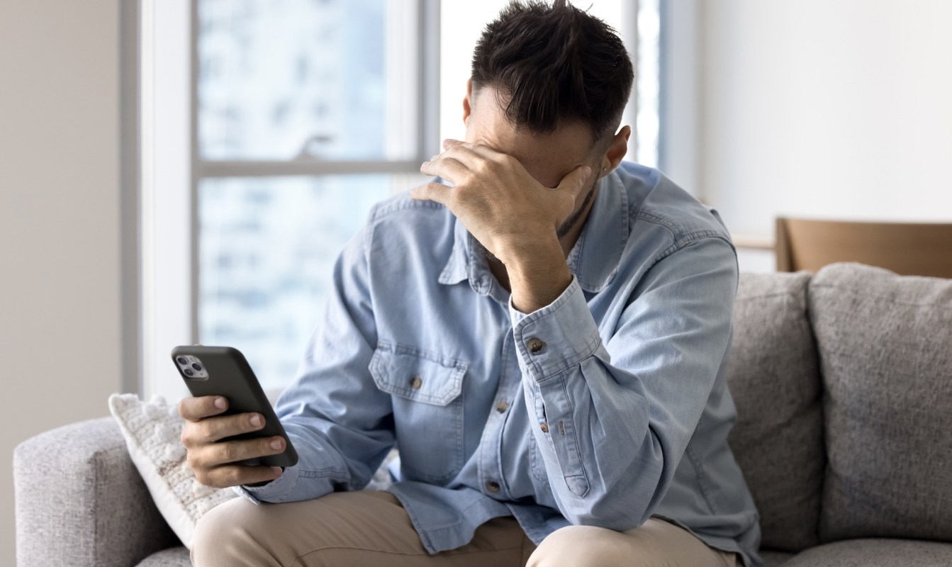 A man sits on a couch, holding a phone and covering his face with one hand, looking distressed.