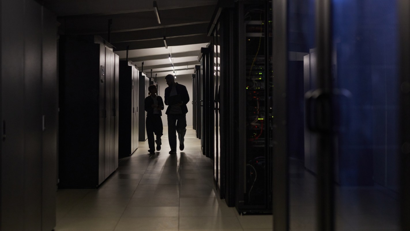 Two individuals walking and talking in a dimly lit server room lined with black server racks.