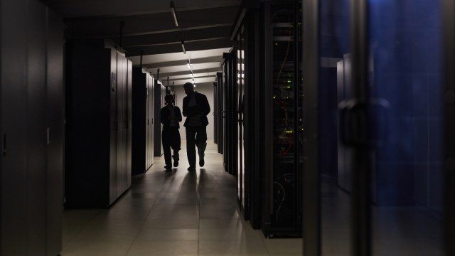 Two individuals walking and talking in a dimly lit server room lined with black server racks.