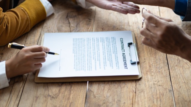A person signs a contract agreement on a wooden table with another individual gesturing.