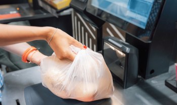A hand is placing a plastic bag of groceries onto a self-checkout station.