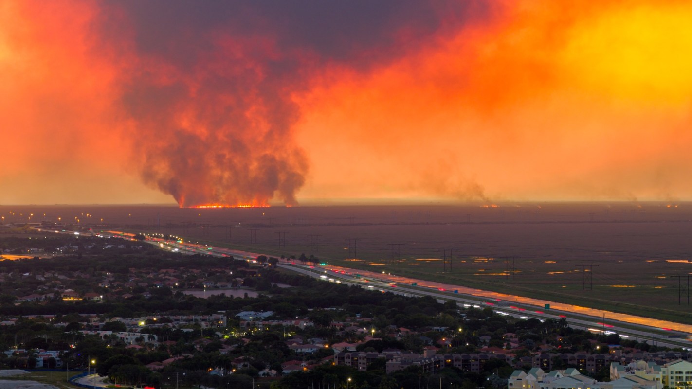 A dramatic sunset over a landscape with smoke rising from wildfires in the distance, and a highway lined with traffic below.