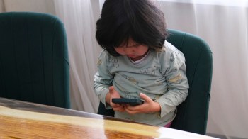 A young child sits at a table, focused on a smartphone, wearing a light-colored shirt with vehicle patterns.
