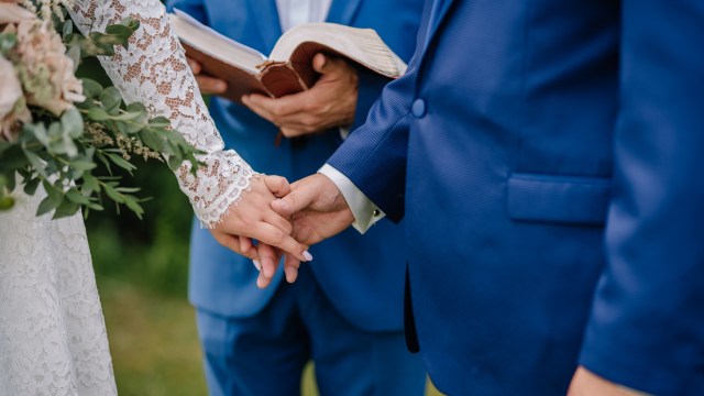 A couple holding hands during a wedding ceremony, with a bouquet and officiant in the background.