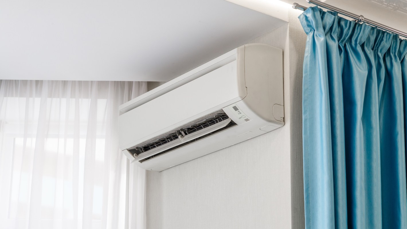 A white mini-split air conditioning unit mounted on a wall beside sheer curtains and blue drapes.