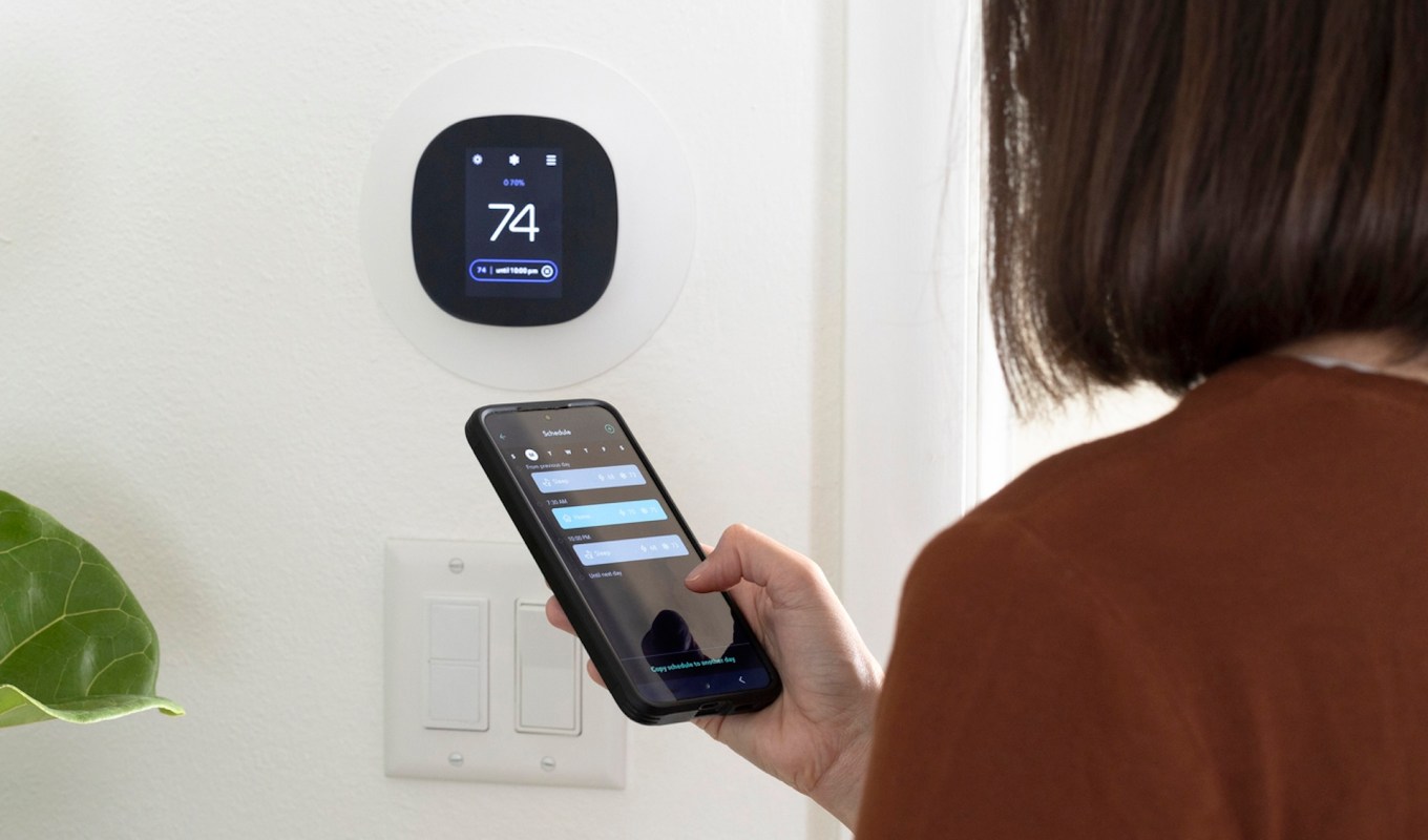 A person adjusts a smart thermostat using a smartphone app while standing next to it indoors.
