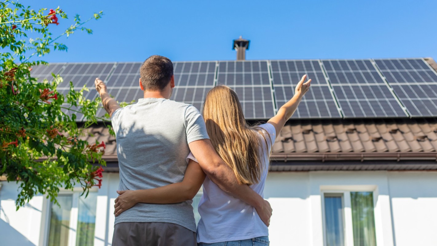 A couple stands together, admiring solar panels on their roof against a clear blue sky.