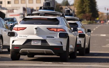 Two white autonomous vehicles from Waymo are stopped at an intersection, showcasing their distinct branding and sensors.