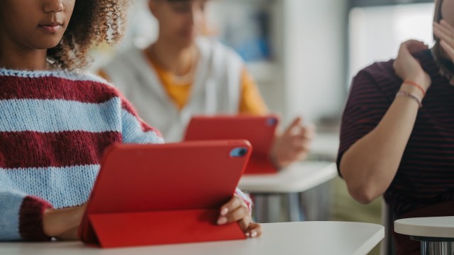 A student in a striped sweater uses a red tablet, while peers in the background are also engaged with their devices.