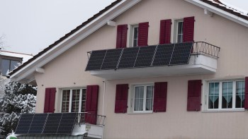 A house with a light-colored façade and maroon shutters featuring solar panels on the balconies, surrounded by snow.