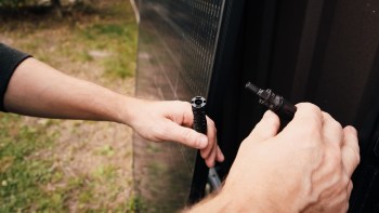 A person connects two black electrical connectors near a solar panel in an outdoor setting.