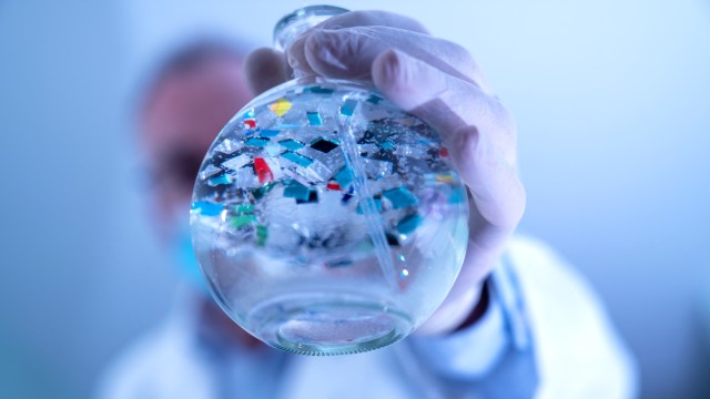A gloved hand holds a round glass flask containing colorful liquid and plastic fragments.