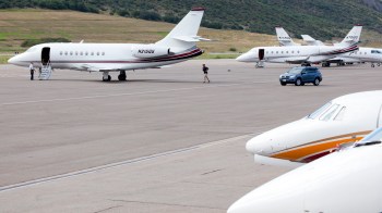 Several private jets parked at an airport with a car driving nearby and a person walking toward one of the planes.