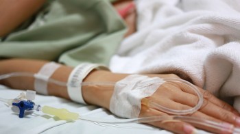 A close-up of a hand with an intravenous (IV) line, resting on a hospital bed.