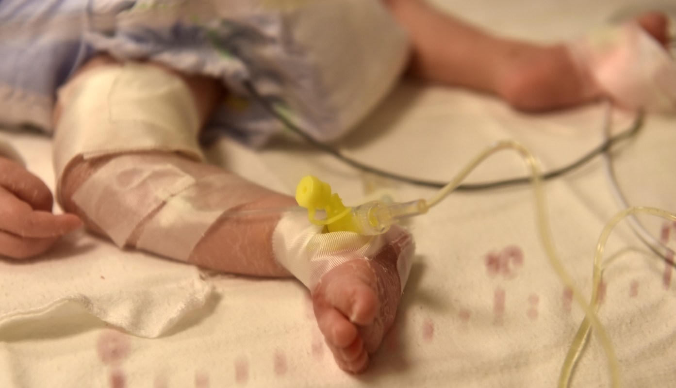 A close-up of a newborn baby's foot with medical tape and an IV catheter attached, lying on a hospital blanket.