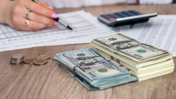 A stack of $100 bills, coins, and financial documents, with a person writing with a pen nearby.