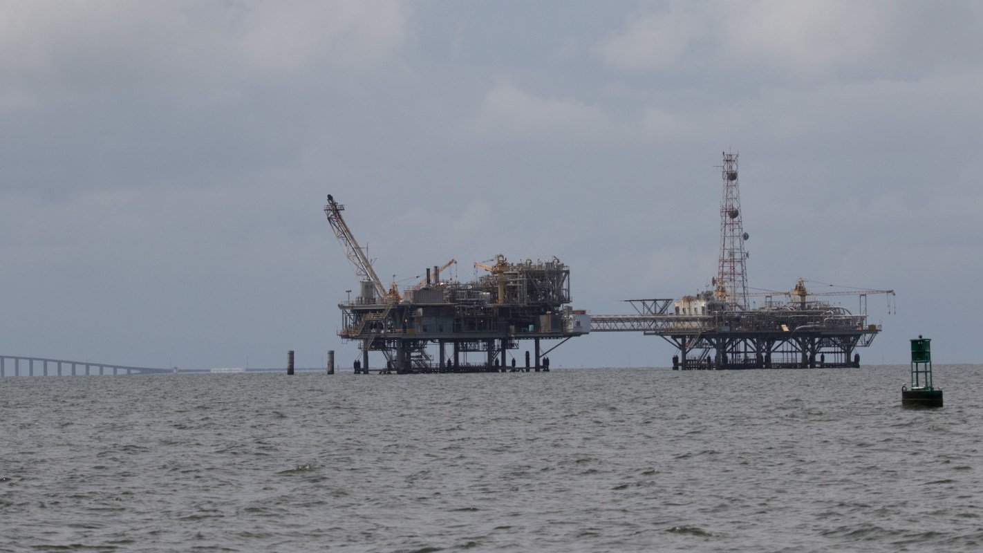 An offshore oil rig operates over a calm sea with a bridge visible in the background under a cloudy sky.