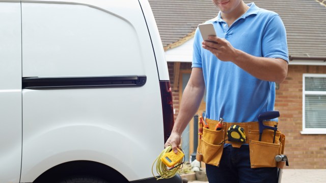 A maintenance worker in a blue shirt checks his phone while holding tools next to a white van.