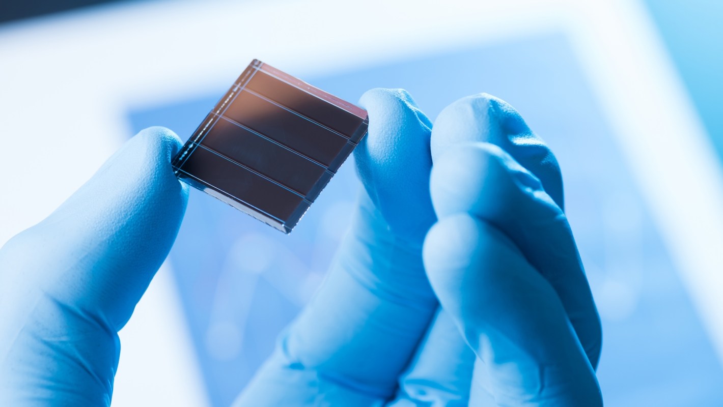A researcher in blue gloves holds a small solar cell against a blurred scientific background.