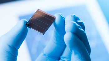 A researcher in blue gloves holds a small solar cell against a blurred scientific background.