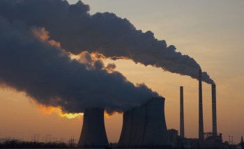 A power plant silhouetted against a sunset, emitting thick smoke from cooling towers and chimneys.