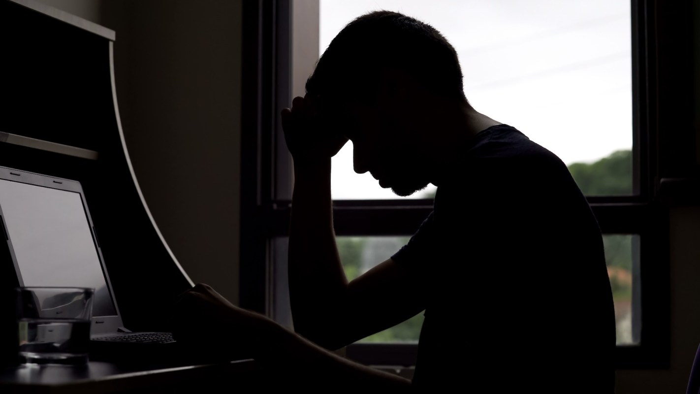 A silhouetted person sits at a desk, resting their forehead on their hand, appearing stressed or burned out.