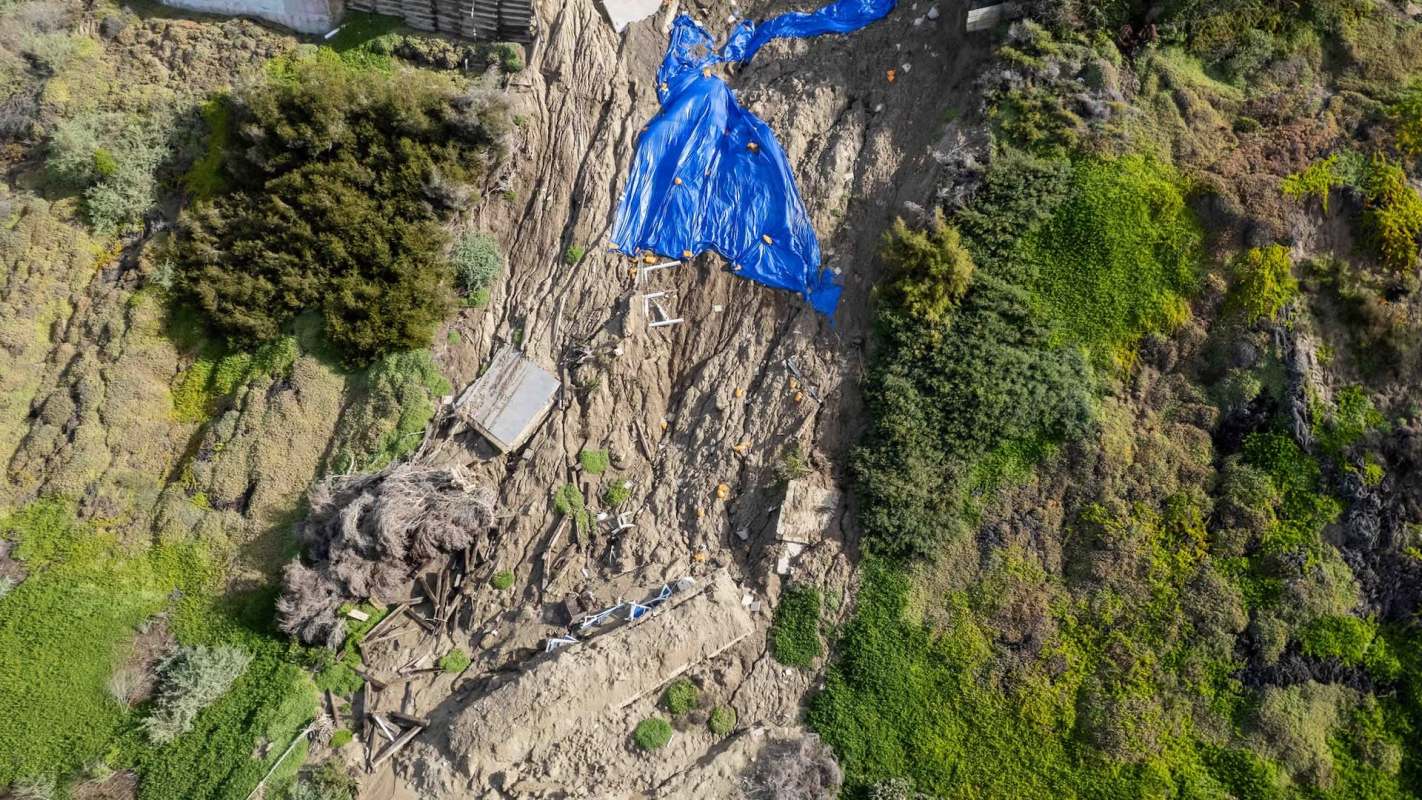 A large section of a hillside has collapsed, exposing soil and debris, covered with blue tarps near residential buildings.