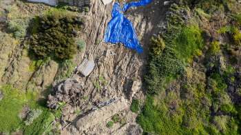 A large section of a hillside has collapsed, exposing soil and debris, covered with blue tarps near residential buildings.