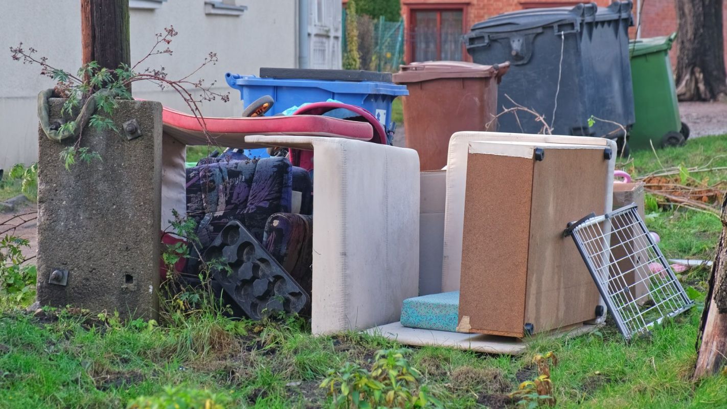A pile of discarded furniture, including chairs and a cabinet, next to trash bins in a grassy area.