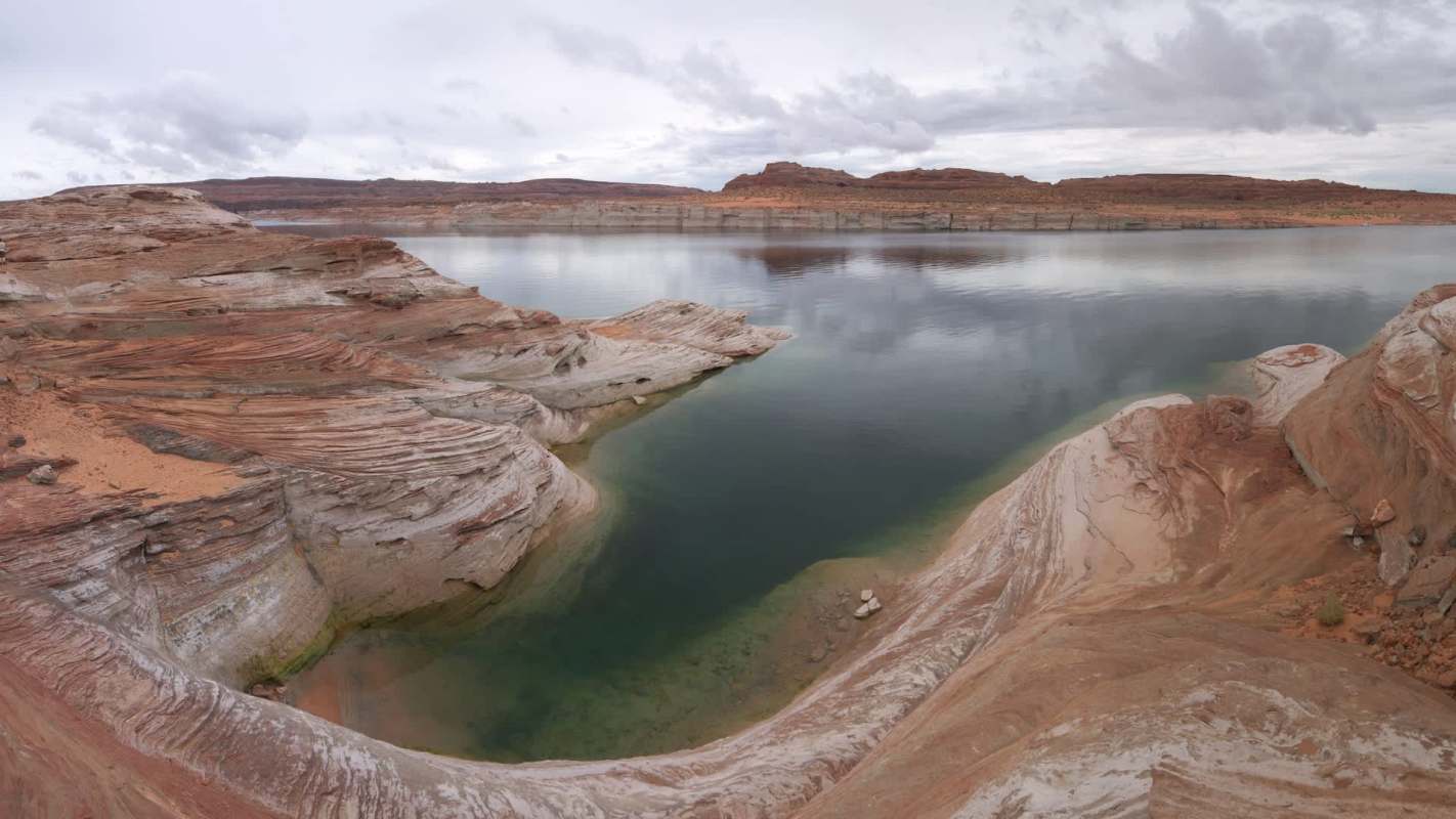 A panoramic view of colorful rock formations surrounding a calm, reflective Lake Powell under a cloudy sky.