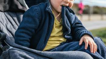 A young child in a blue jacket bites a piece of his clothing while seated in a stroller on a sunny day.
