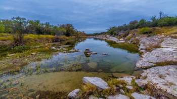 A tranquil river scene surrounded by greenery and rocky banks under a cloudy sky.