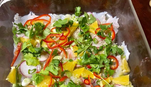 A vibrant dish featuring raw fish topped with herbs, citrus, sliced chili, and radishes in a clear glass dish.