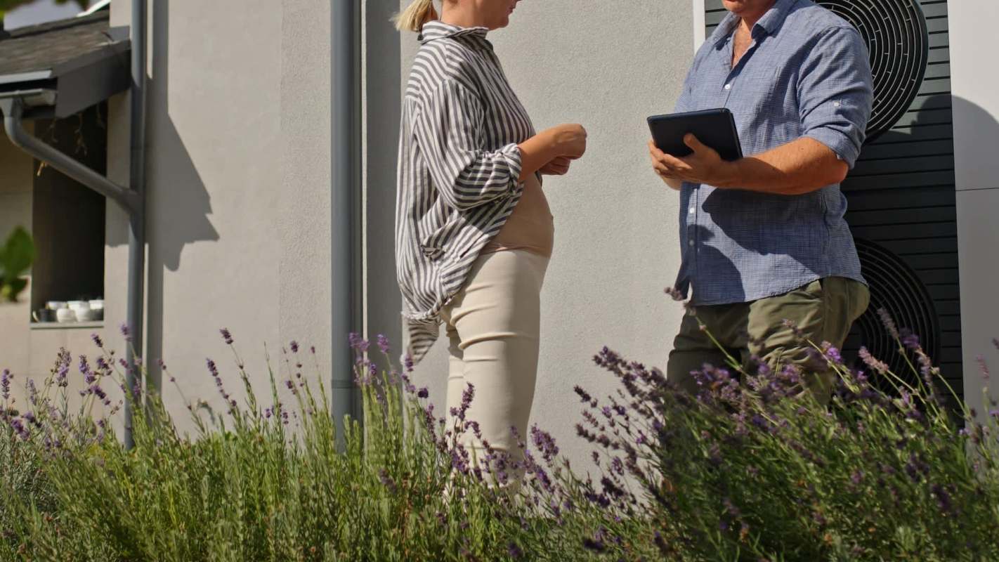 A woman in a striped shirt talks with a man in a blue shirt with a tablet near a heat pump outside of a house.