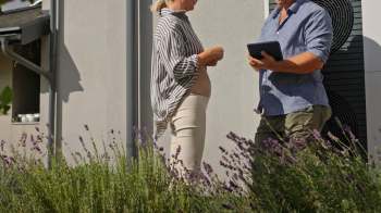 A woman in a striped shirt talks with a man in a blue shirt with a tablet near a heat pump outside of a house.