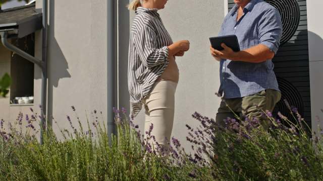 A woman in a striped shirt talks with a man in a blue shirt with a tablet near a heat pump outside of a house.