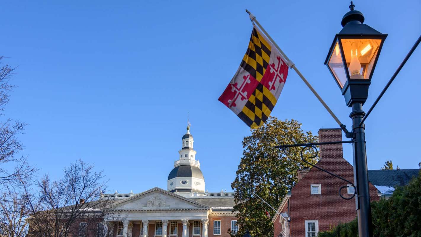 A Maryland flag flutters in front of a historic building under a clear blue sky.