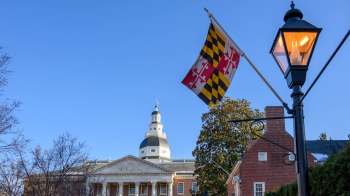 A Maryland flag flutters in front of a historic building under a clear blue sky.