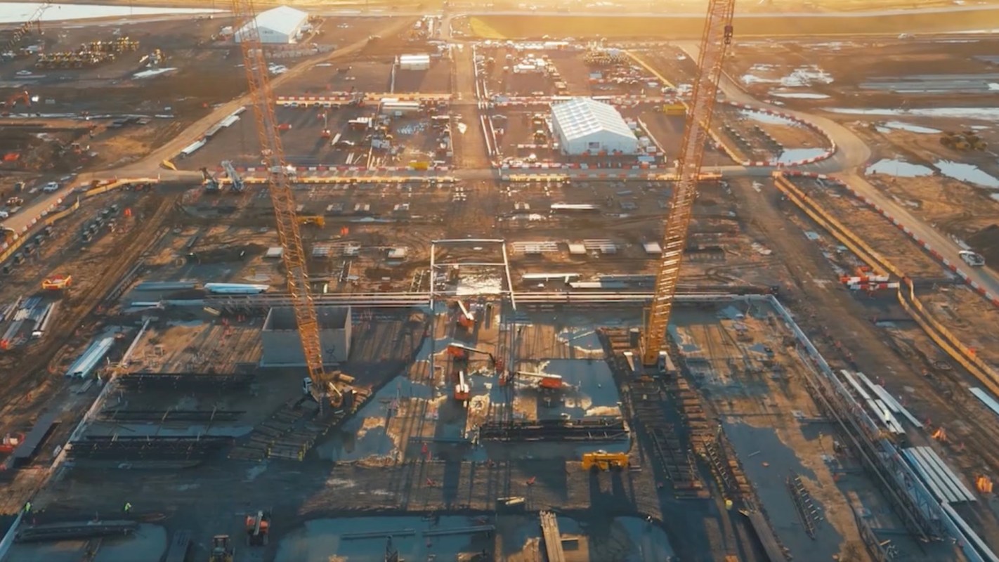 An aerial view of a construction site with cranes, equipment, and materials scattered across a muddy terrain at sunset.