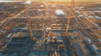 An aerial view of a construction site with cranes, equipment, and materials scattered across a muddy terrain at sunset.