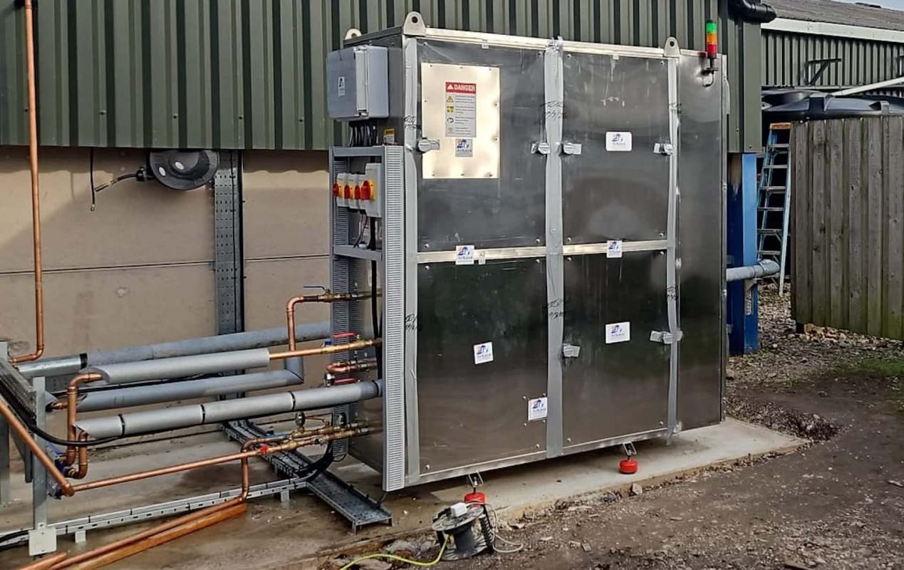 A metallic industrial unit surrounded by pipes, mounted on a concrete base outside a building.