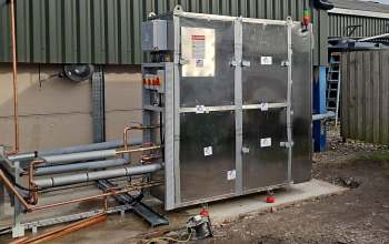 A metallic industrial unit surrounded by pipes, mounted on a concrete base outside a building.