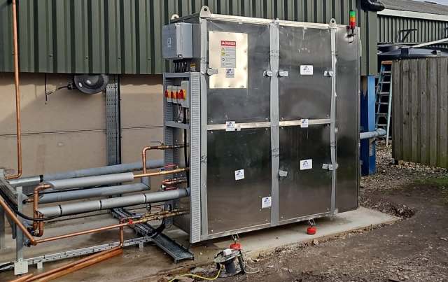 A metallic industrial unit surrounded by pipes, mounted on a concrete base outside a building.