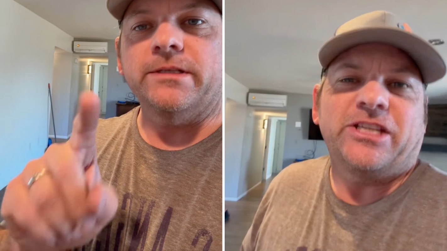 A man with a baseball cap gestures to a mini-split unit on a wall behind him while speaking in a room.