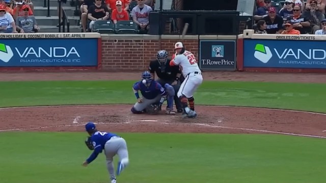 A baseball player prepares to hit as the pitcher throws a ball during a game with the umpire and fans in the background.