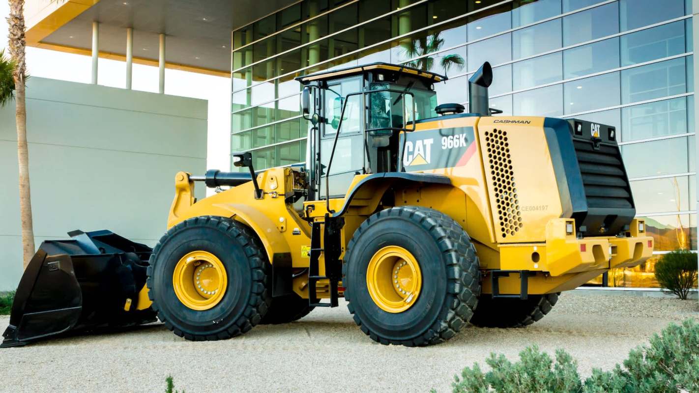 A yellow Caterpillar 966K wheel loader is parked outside a modern building with glass reflections and landscaping.
