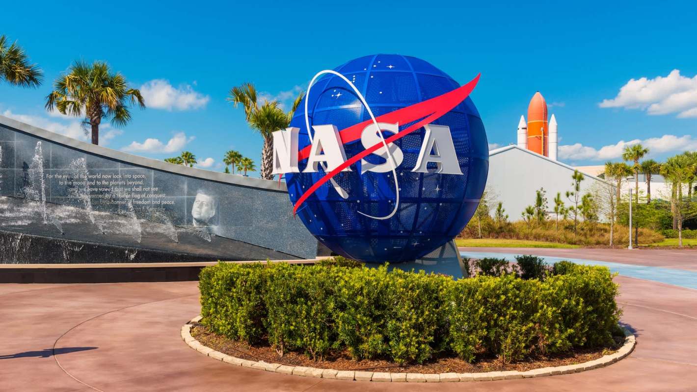 A large blue NASA globe sign with a space shuttle in the background, surrounded by palms and greenery.