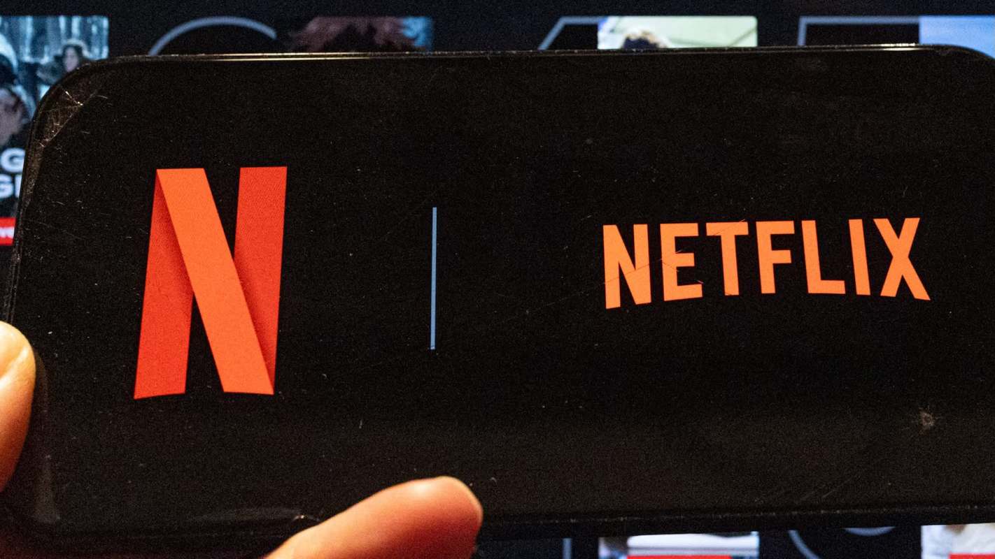 A hand holding a smartphone with the Netflix logo in front of a screen.