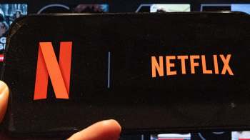 A hand holding a smartphone with the Netflix logo in front of a screen.
